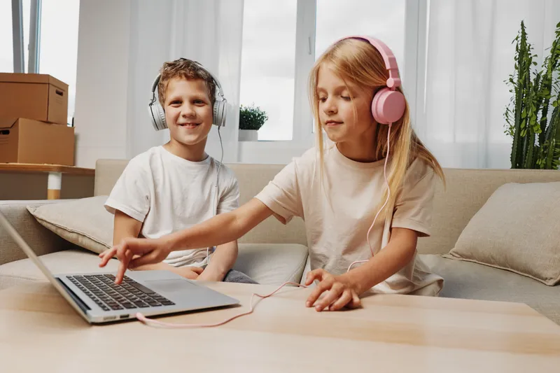 Two children wearing headphones enjoy music together, one using laptop, the other smiling, in cozy living room with natural light and plants visible.