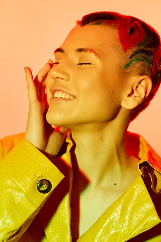 Smiling woman with colorful dyed hair and yellow jacket enjoys warm light. Bright makeup, positive mood, close-up portrait with vibrant orange background.
