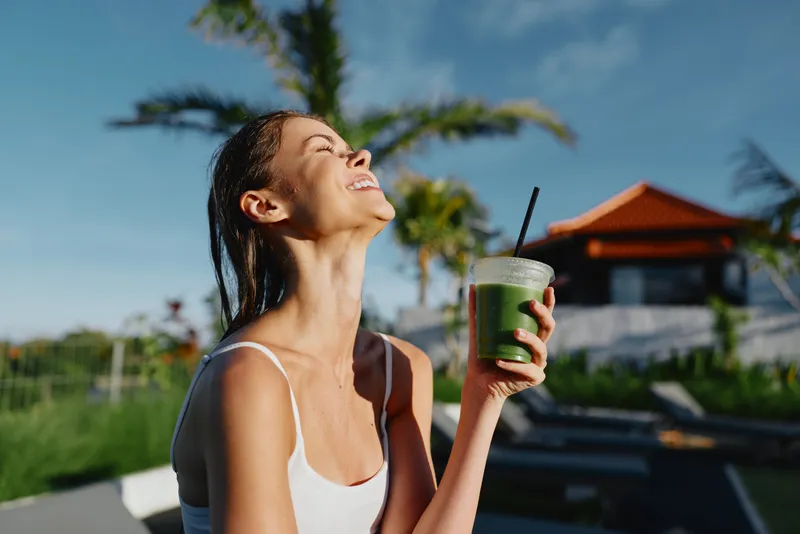 Joyful woman enjoys a refreshing green smoothie outdoors, by a pool under clear skies, radiating health, leisure, and happiness in a casual summer moment as sunlight glows.