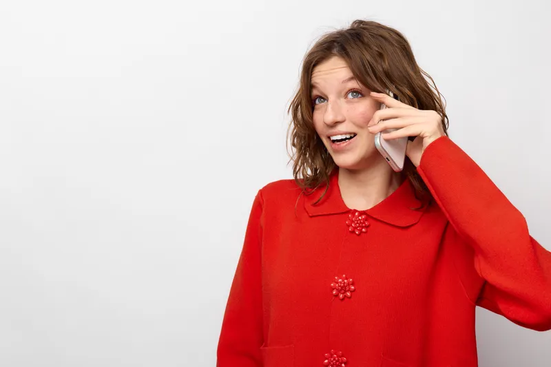 Young woman in red sweater talking on smartphone with happy expression isolated on white background. Concept of communication, technology, and casual lifestyle.