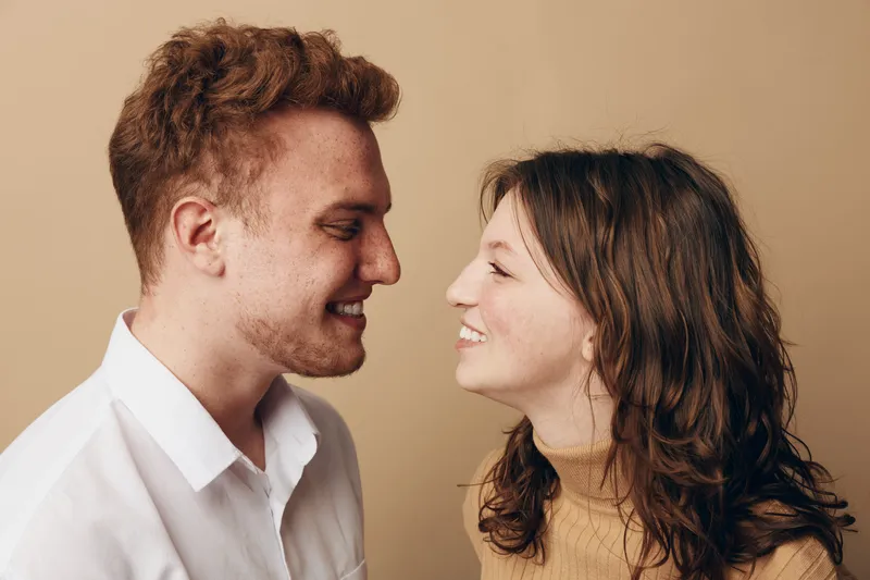 Smiling young couple face to face close up, looking at each other with happy expressions. Intimate moment of love and connection between man and woman on beige background.