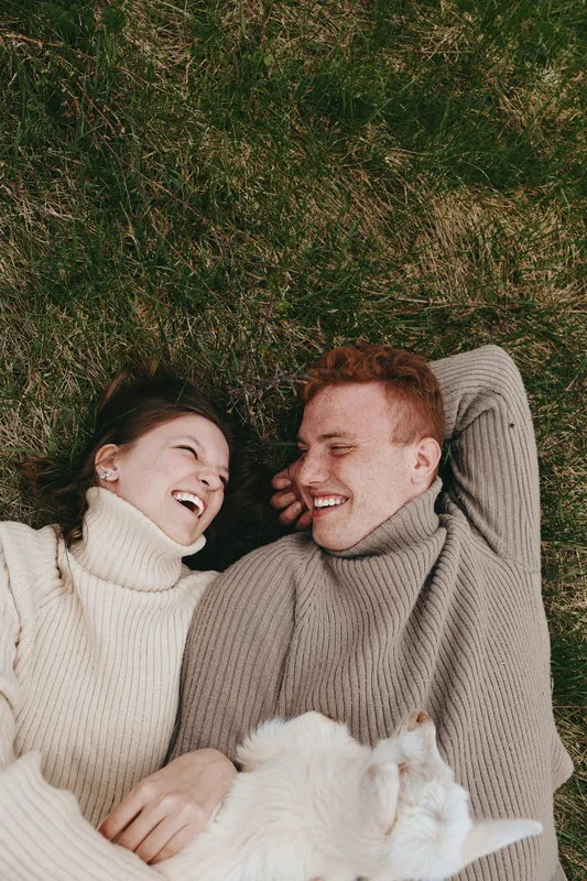Cheerful couple outdoors, lying on grass, laughing in warm sweaters, sunlit moment, relaxed posture, soft breeze, natural colors, dog nearby enjoying calm companionship