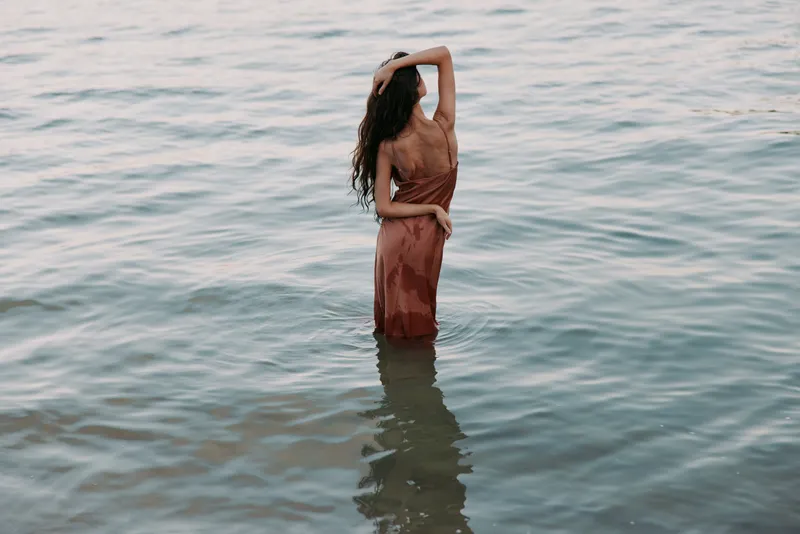 Woman stands in calm sea, wearing a flowing bronze dress, back to camera, soft light creates a reflective surface and graceful silhouette, conveying calm and timeless summer mood.