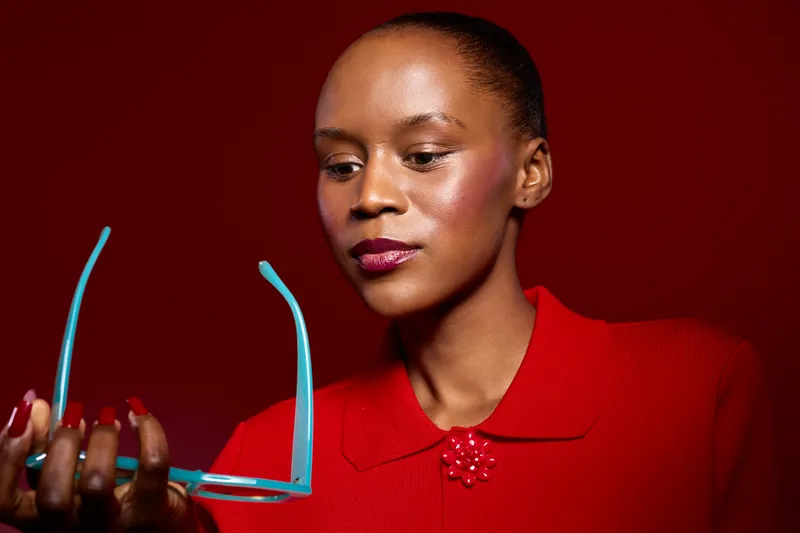 Portrait of an African woman with dark skin wearing a red blouse holding blue eyeglasses. Studio shot on red background with soft lighting. Concept for fashion, beauty, and accessories.