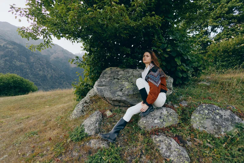 Woman sitting on rocks under a tree by mountains, wearing a patterned sweater and boots in an autumn meadow, portrait-style outdoor scene conveying cozy, peaceful nature escape.