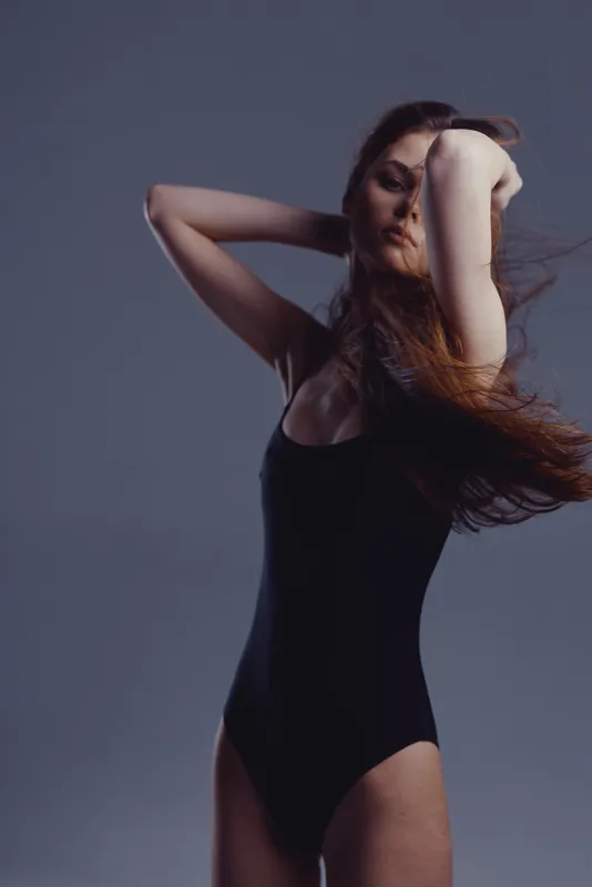 elegant woman in black bodysuit posing with closed eyes against gray background in studio lighting
