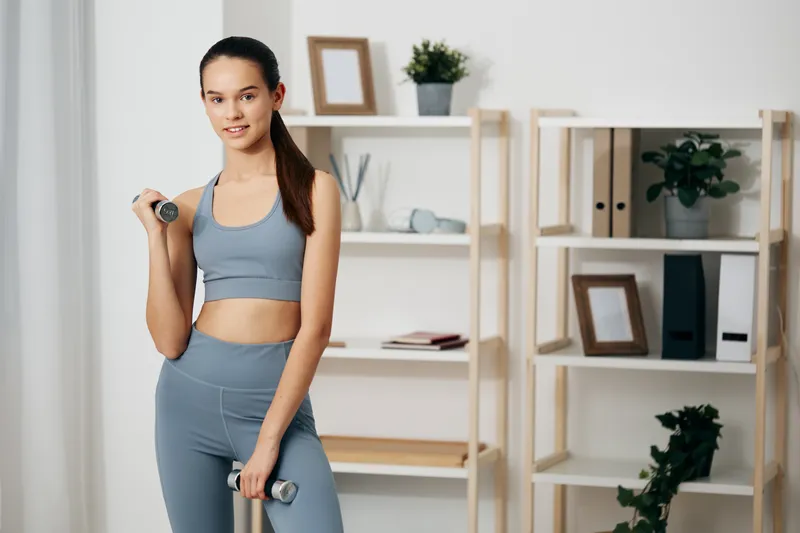 A fit woman in blue athletic wear stands with dumbbells in a contemporary home gym, ready for a workout session. Modern interior with shelves and plants highlighting wellness, fitness, strength and