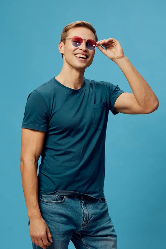 Smiling man wearing casual blue t-shirt and red sunglasses pose with hand adjusting glasses against blue background. Studio portrait of happy cheerful young Caucasian male in summer clothing. People l