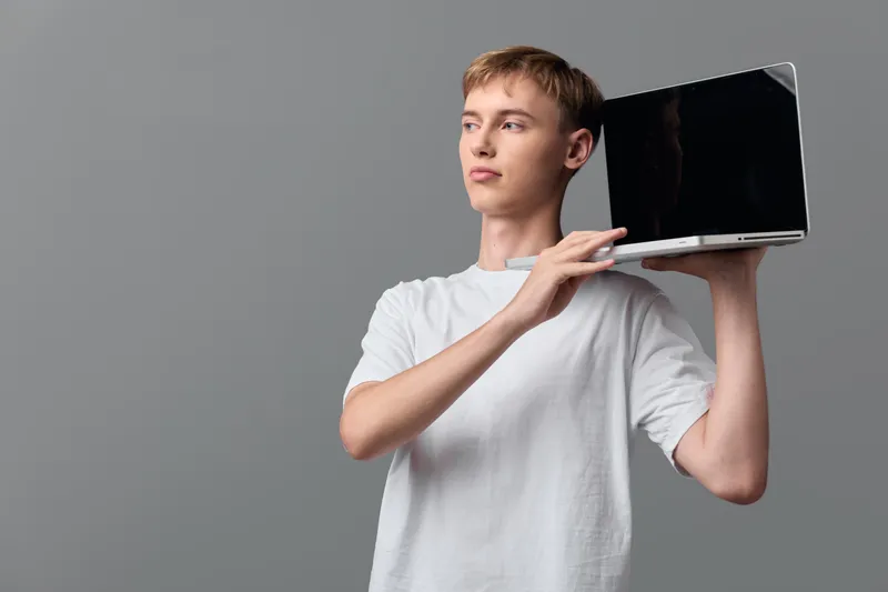 Casual man holding a laptop by the edge against a neutral gray background, highlighting portable technology use, everyday digital work, and hands on learning