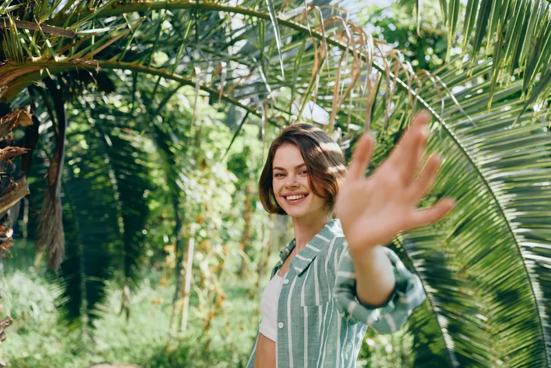 Cheerful portrait of a smiling woman in a lush tropical garden, reaching toward the camera with a friendly gesture amid greenery.