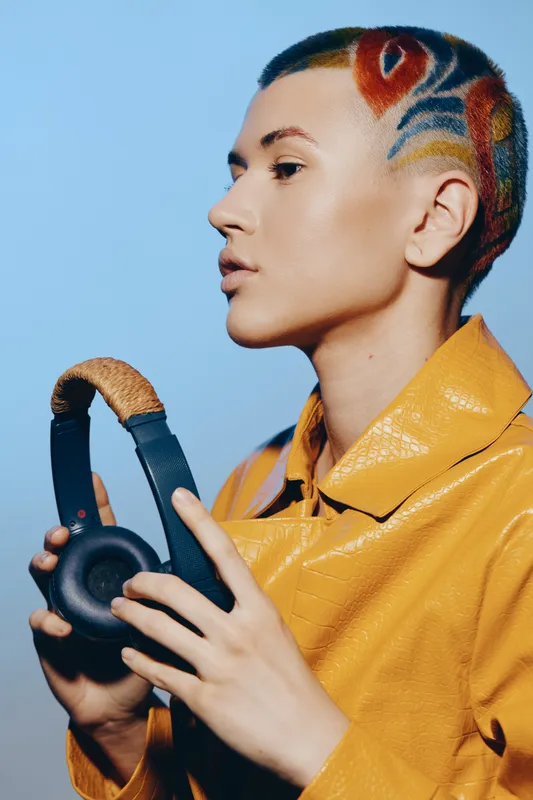 young woman with artistic short hairstyle holding black headphones, wearing yellow textured jacket, blue background, modern style, creative urban portrait