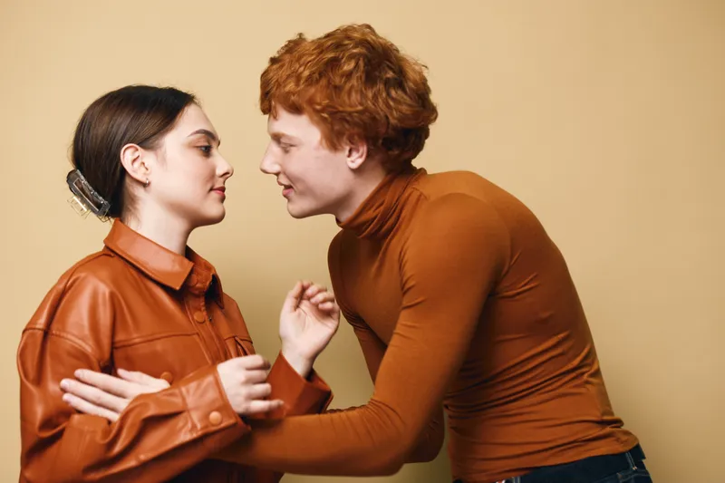 Young couple with red hair and brown leather jacket posing close in studio, intimate moment with eye contact and soft expression on warm beige background.