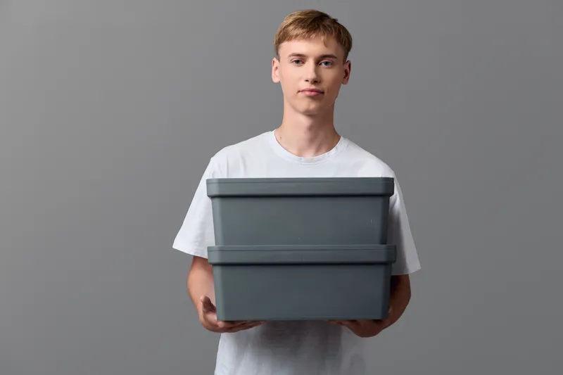 Man in a plain tee holds two gray storage boxes, demonstrating home organization and light moving, with emphasis on packaging, storage solutions, and orderly transfer concepts