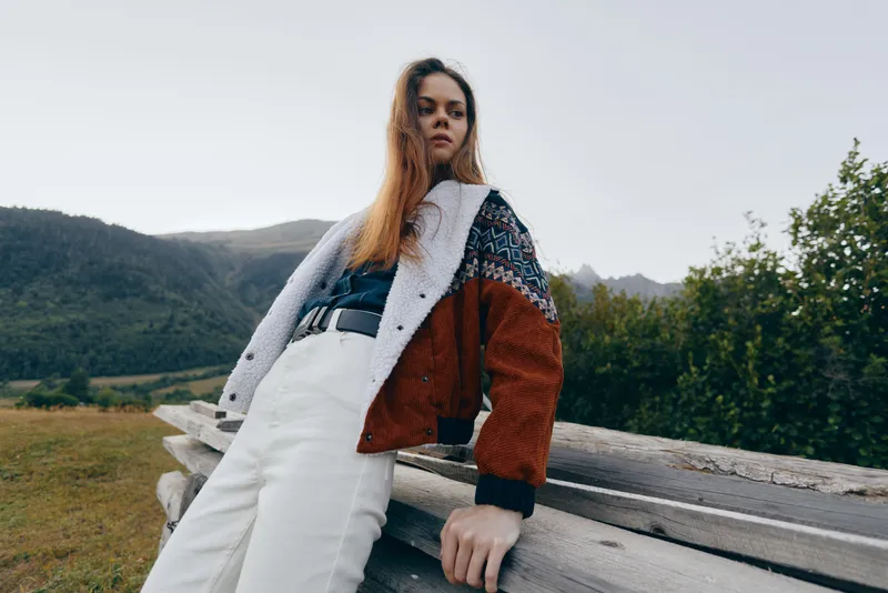Woman portrait outdoors in countryside by wooden fence, wearing autumn jacket and white pants, fashion model posing against mountains for seasonal style, travel and relaxed lifestyle vibe.