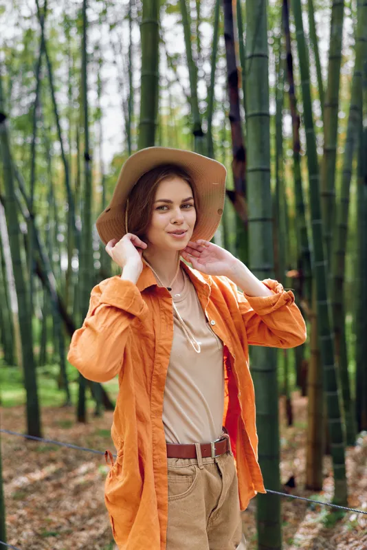 Woman portrait in bamboo forest wearing a straw hat and orange jacket, smiling at camera. Outdoor travel lifestyle, casual outfit and serene nature walk with green foliage background.