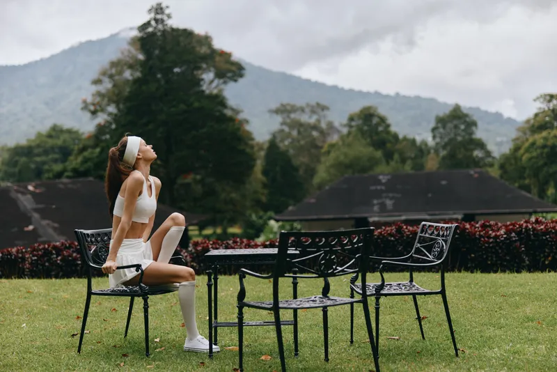 Young athletic woman in white sportswear sitting outdoors on a chair, showcasing strength and confidence amidst a serene landscape of greenery and mountains