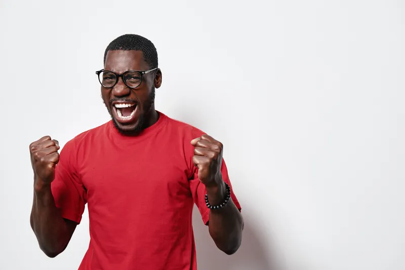 African American man with dark skin wearing glasses and red t shirt celebrating success with clenched fists and open mouth on white background. Excited, happy and energetic expression.