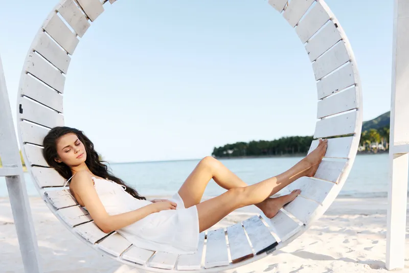 Portrait of a relaxed woman in a white dress seated in a circular beach swing by the shore, sunlight, blue sea, and sandy beach creating a serene summer scene.