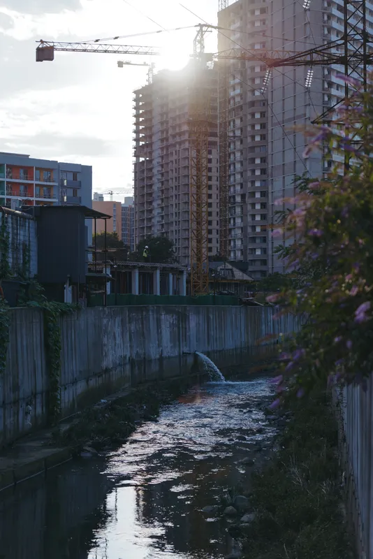 Construction site with highrise buildings beside a river canal and tower crane at sunset, urban development scene with concrete embankment and waterway reflecting sun flare and dusk light.