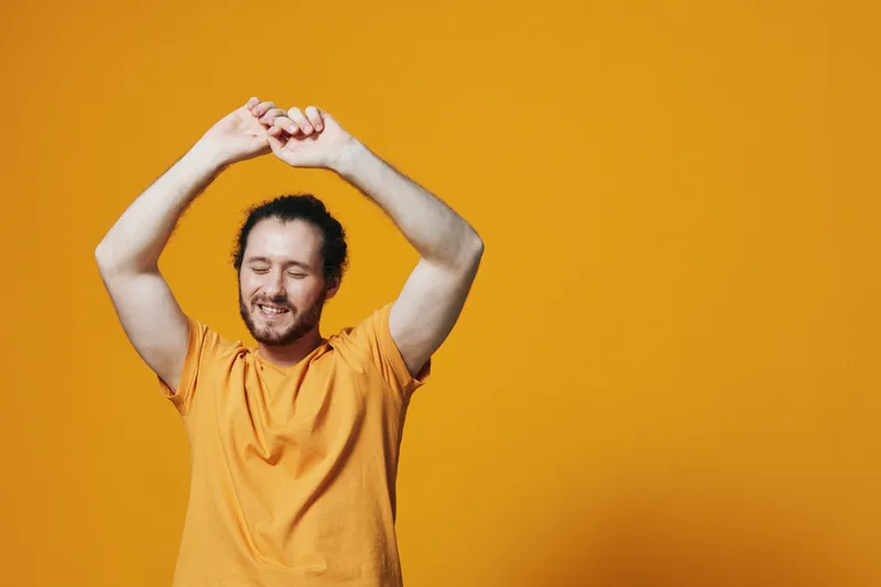 Happy young man stretching with a joyful expression against a bright yellow background, wearing a casual orange t shirt, conveying relaxation and positivity