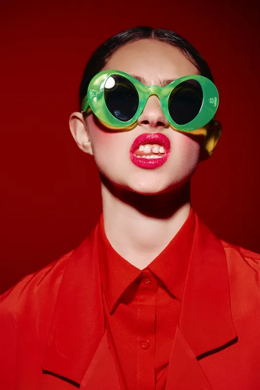 Young woman in bright red outfit wears vivid green oversized sunglasses and glossy red lipstick against deep red background. Creative fashion portrait expressing bold and edgy beauty style.