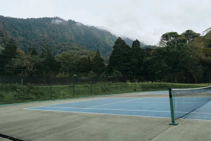 Serene outdoor tennis court surrounded by lush green mountains under a cloudy sky, perfect for sports enthusiasts seeking tranquility and natural beauty
