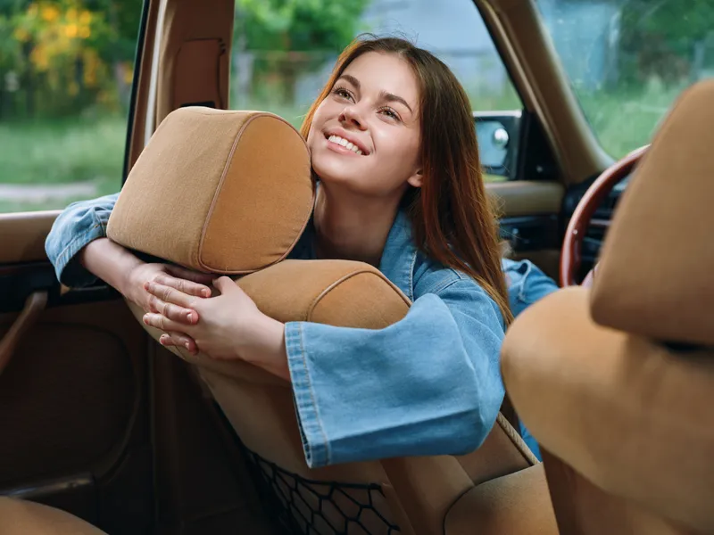 Young woman enjoying a moment of happiness in a car, wearing a denim jacket, with an engaging expression, set against a soft green background.