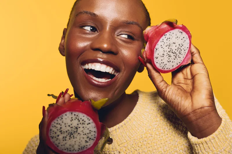 Happy African woman with dark skin holding two halves of dragon fruit near her face, smiling and looking away. Bright yellow background and casual sweater create vibrant, fresh mood.