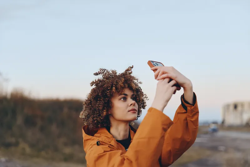 Woman curly hair orange jacket holding phone outdoors in bright daylight, lifestyle travel mood, pose while preparing a selfie, open sky and blurred street background, distant buildings