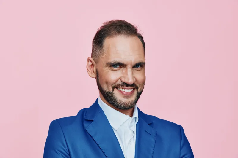 A confident male business professional stands against a solid pink background, smiling warmly. He exudes optimism and focus, suggesting a capable leader ready to tackle challenging projects.