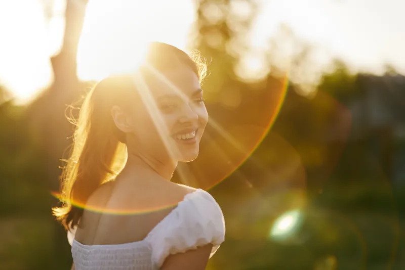 Smiling young woman enjoying a sunny day outdoors, wearing a white summer dress, radiating joy and positivity in a vibrant natural setting