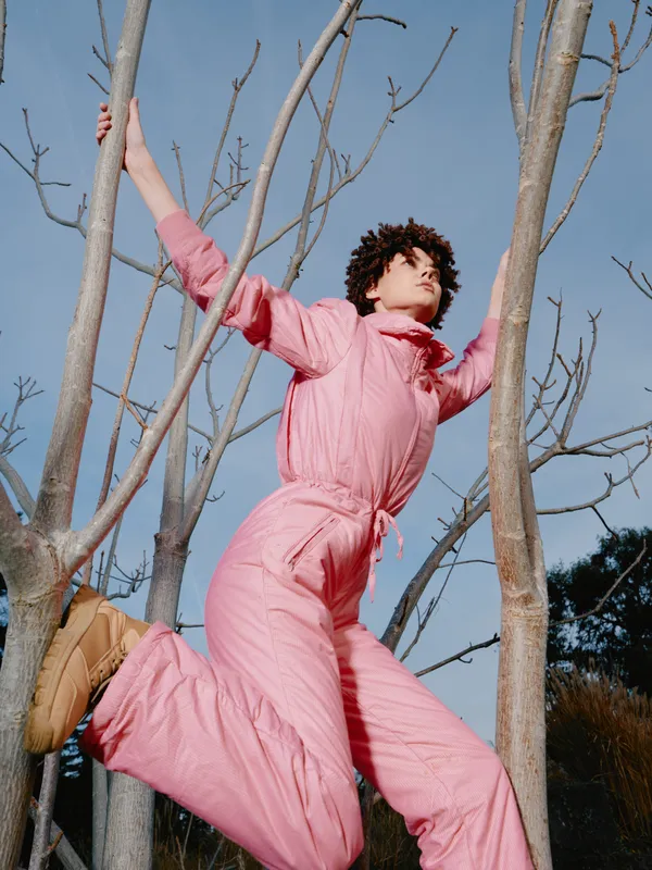 Person in pink jumpsuit leaps among bare trees outdoors, showcasing athletic energy and dynamic movement under a bright clear sky.