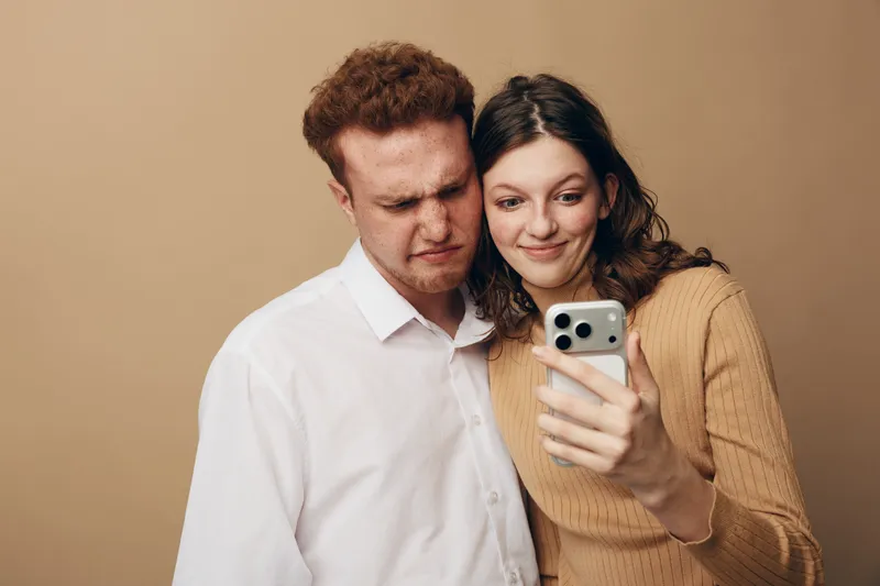 Young couple takes a selfie together, woman smiling and man making a confused face. Casual clothing and neutral background create a relaxed, candid moment of shared technology use.