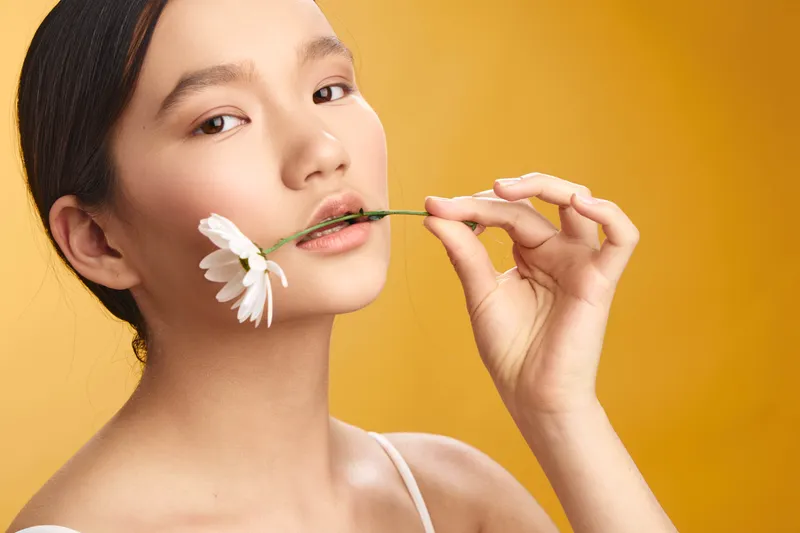 Asian woman holds a delicate daisy near her lips, set against a warm yellow backdrop, creating a calm studio portrait that exudes creative charm and poised elegance.