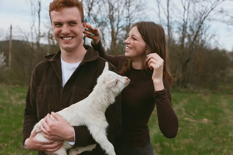 Young couple enjoys spending time outdoors with a white baby goat in a green field, sharing joyful moments and smiling naturally in casual autumn clothing.