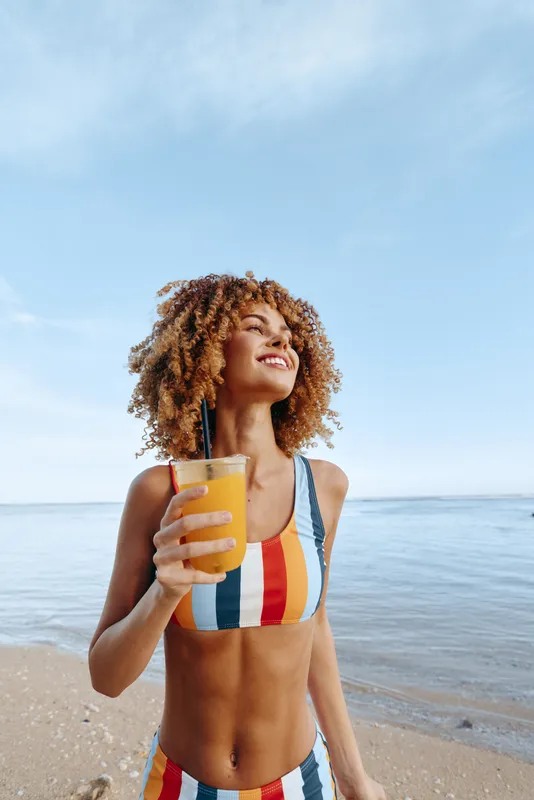 Young woman with curly hair wearing a colorful striped swimsuit enjoys a fresh orange juice on a sandy beach under a clear blue sky with a joyful smile.