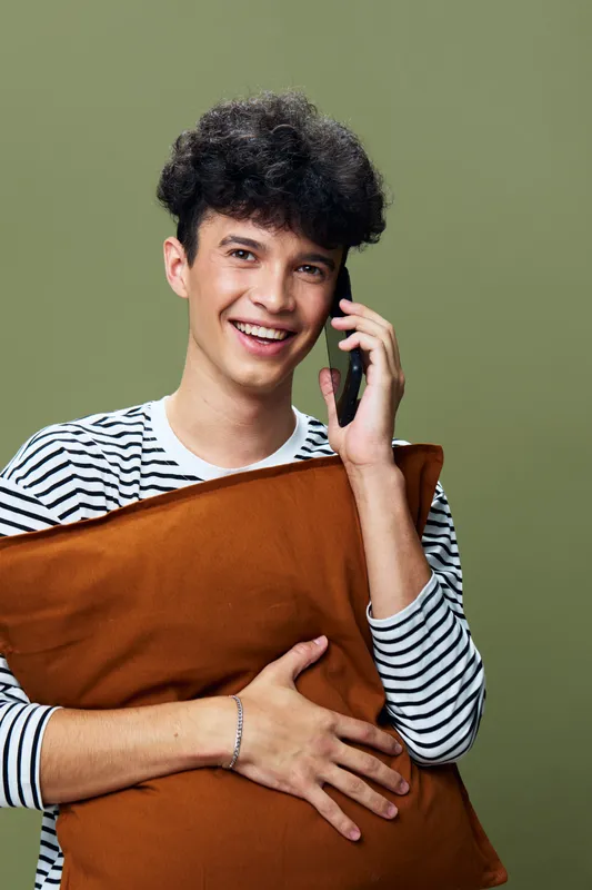 smiling man holding brown pillow talking on phone wearing striped shirt with green background, young man communicating happily indoors