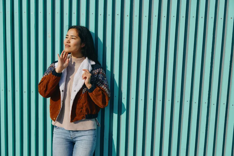Street fashion woman leaning against a turquoise wall in a textured jacket and denim, exuding casual confidence and urban vibe under bright sun, ideal for contemporary lifestyle campaign