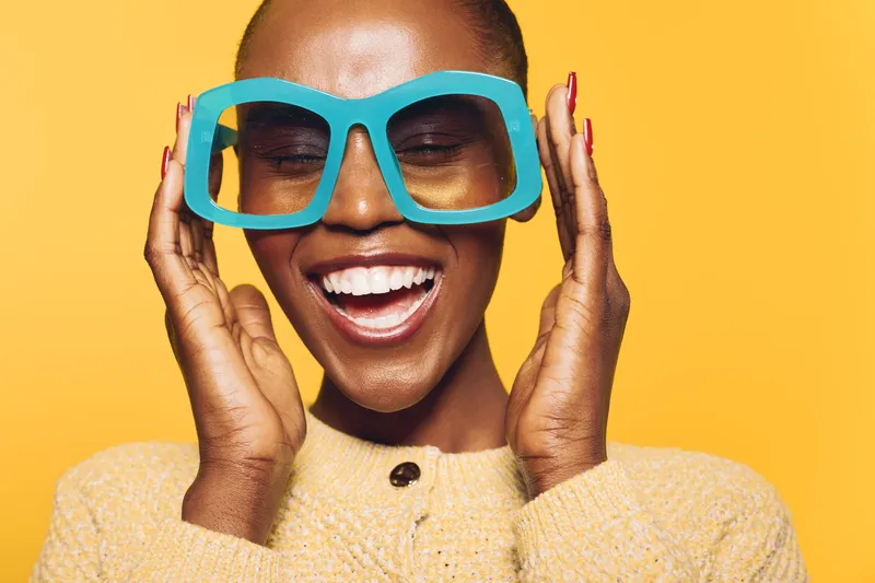 Happy African woman with dark skin wearing oversized blue glasses and yellow sweater smiles with closed eyes on bright yellow background. Concept of joy, fashion, and vibrant lifestyle.
