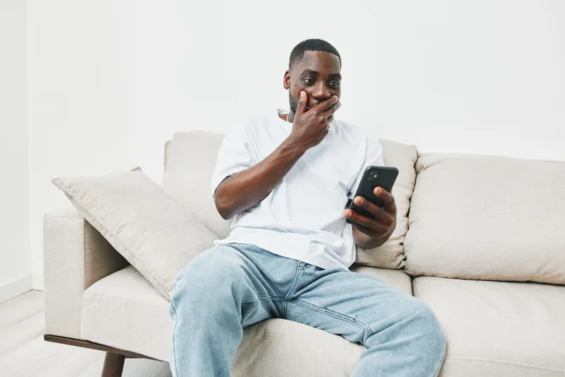 Shocked young African American man looking at his smartphone while sitting on a couch in a modern living room