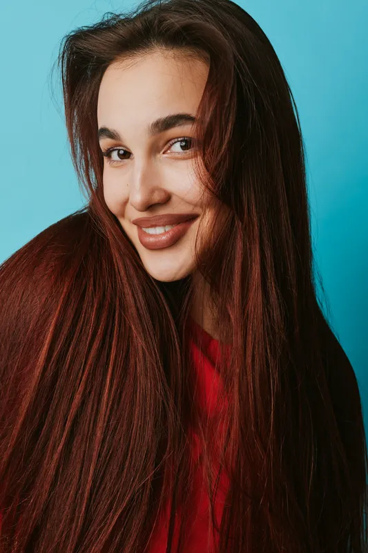 Young woman with long brown hair smiling and looking at camera on blue background. Portrait of happy female with natural makeup and casual style for beauty and lifestyle.