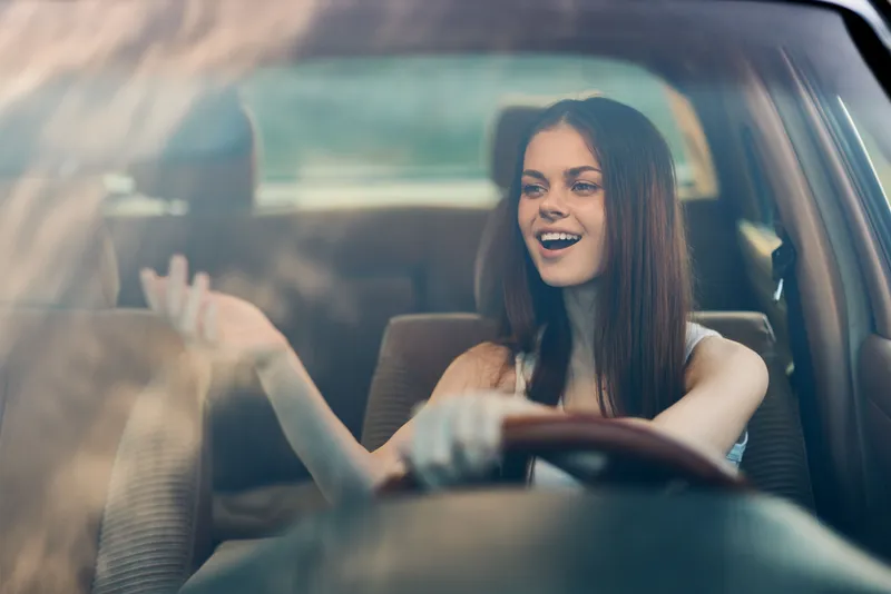 Happy young woman enjoying a road trip, singing while driving in a car, with natural lighting highlighting her joyful expression and casual attire.