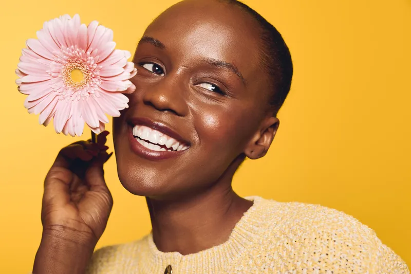 Smiling black woman with dark skin holds pink flower near face on yellow background. Close-up portrait with natural makeup and cozy sweater. Concept of beauty, happiness, and skincare.