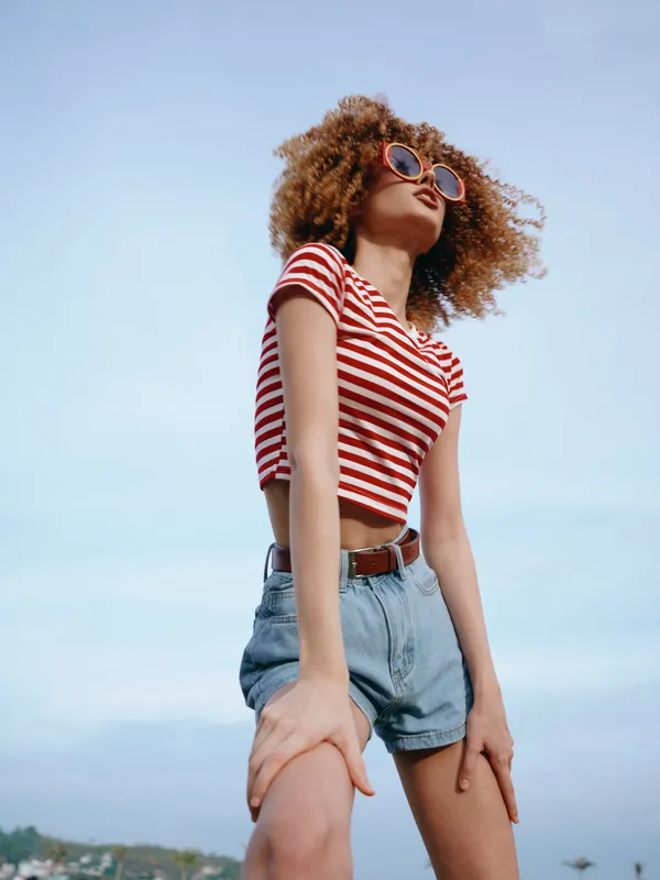 Young woman with curly hair in a red striped crop top and denim shorts stands against a bright blue sky, exuding confidence, freedom, and carefree summer vibes.