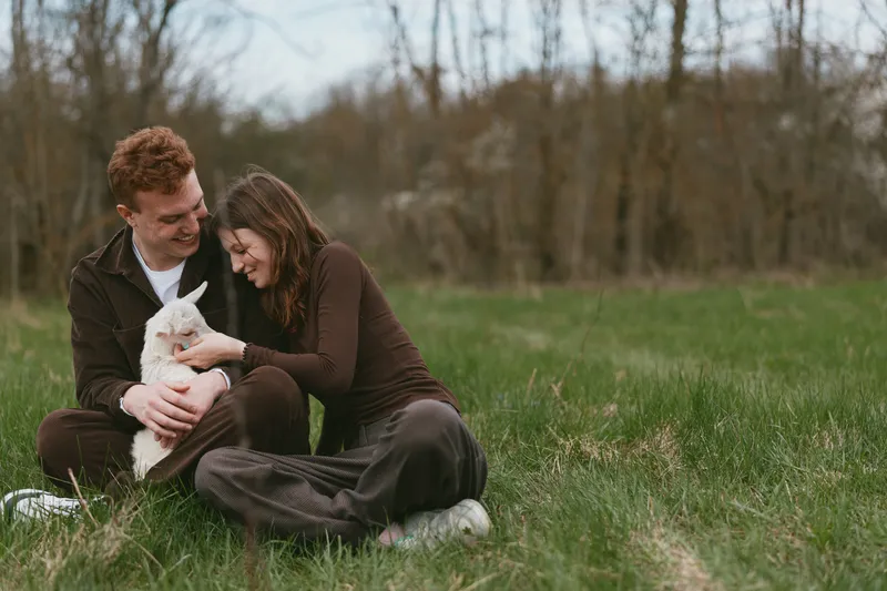 A couple sits on the grass outdoors, cuddling a small goat kid in a sunny meadow, capturing rustic family life, simple pleasures, and gentle countryside moments of care.
