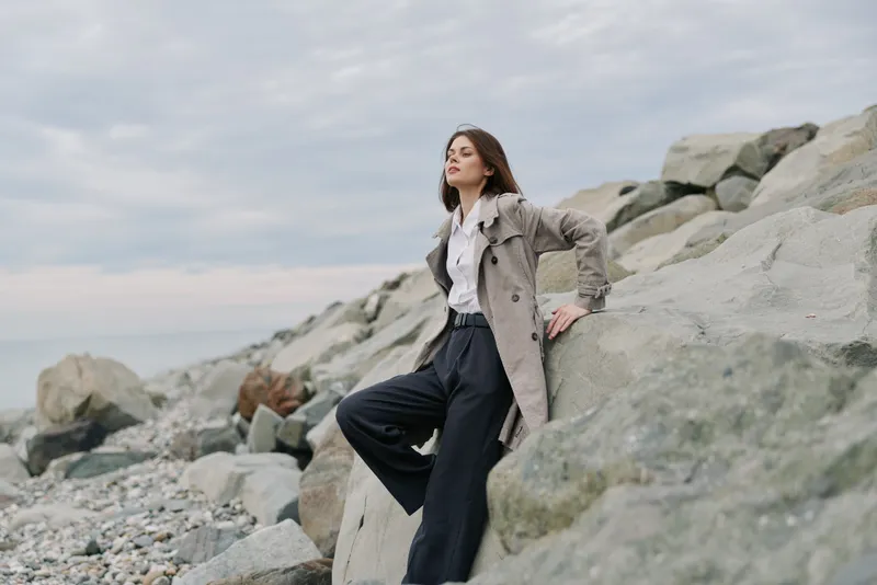 Young woman in a long coat poses confidently on rocky shore. Overcast sky and natural stones create a serene outdoor atmosphere, concept for fashion, nature, and lifestyle photography.