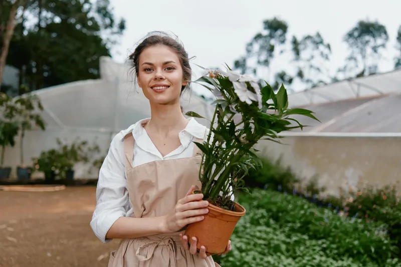 Young woman in an apron happily holding a potted plant in a greenhouse, showcasing a connection with nature and sustainable gardening
