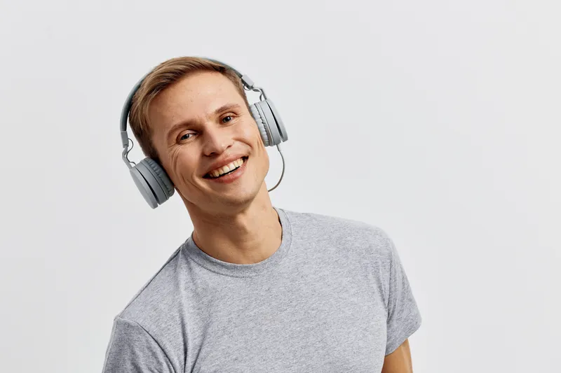 Smiling young man with headphones wearing casual grey t-shirt isolated on plain white background. Studio portrait of happy male enjoying music, cheerful expression, relaxed mood, positive energy, life