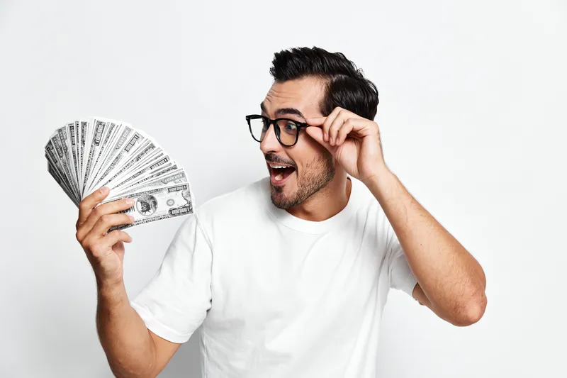Excited smiling young man wearing glasses holds a fan of cash while looking at it, displaying happiness and surprise against a solid white background.