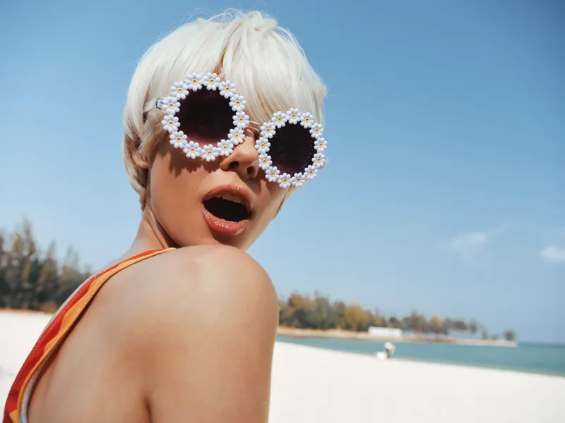 Close up of a stylish beachgoer wearing playful flower sunglasses, sunlit skin, and a surprised expression, capturing carefree summer vibes and bold fashion on a bright seaside day.